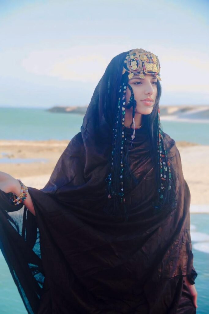 Portrait artistique de Manal Cherki se tenant pieds nus sur le seuil d'une porte, entre deux battants en bois sculpté. Elle porte une robe fluide aux motifs délicats, les bras gracieusement croisés au-dessus de la tête devant un voilage blanc. Une bougie au premier plan apporte une touche de lumière chaleureuse et introspective.