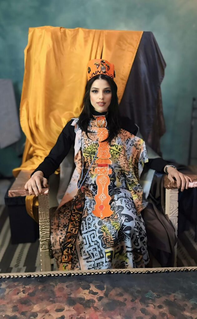 Portrait artistique de Manal Cherki se tenant pieds nus sur le seuil d'une porte, entre deux battants en bois sculpté. Elle porte une robe fluide aux motifs délicats, les bras gracieusement croisés au-dessus de la tête devant un voilage blanc. Une bougie au premier plan apporte une touche de lumière chaleureuse et introspective.