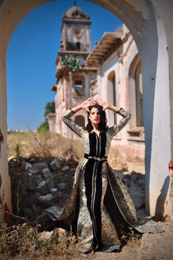 Portrait artistique de Manal Cherki se tenant pieds nus sur le seuil d'une porte, entre deux battants en bois sculpté. Elle porte une robe fluide aux motifs délicats, les bras gracieusement croisés au-dessus de la tête devant un voilage blanc. Une bougie au premier plan apporte une touche de lumière chaleureuse et introspective.