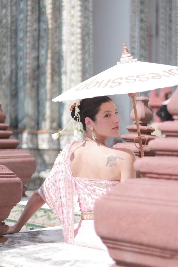 Accueil 3 Manal Cherki de dos, vêtue d'un habit traditionnel orné de perles, tournant le regard avec douceur sous une ombrelle blanche au cœur d'un site patrimonial.
