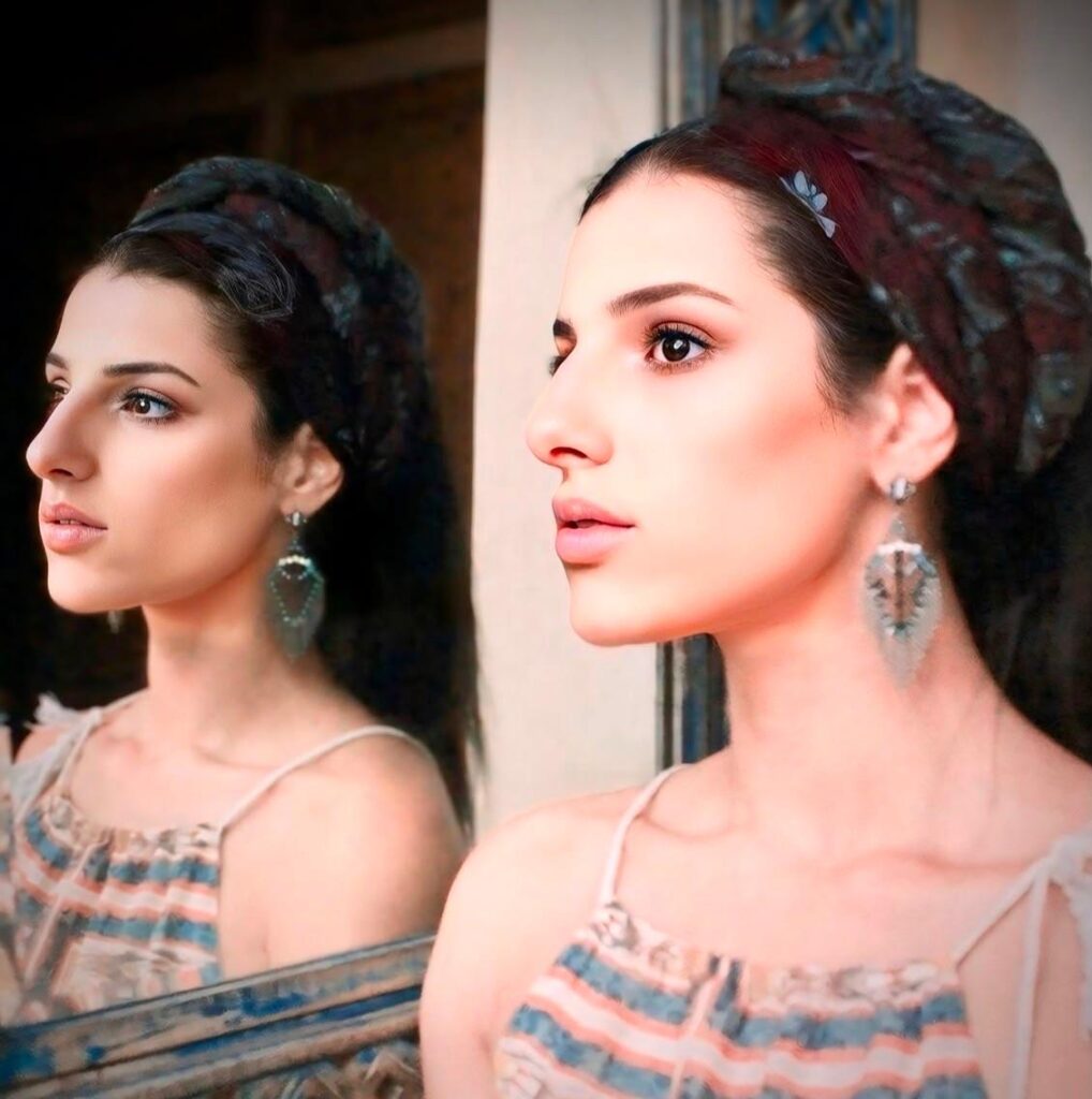 Accueil 1 Portrait artistique de Manal Cherki regardant vers l'horizon, avec son reflet paisible dans un miroir, portant d'élégantes boucles d'oreilles et une coiffe texturée.
